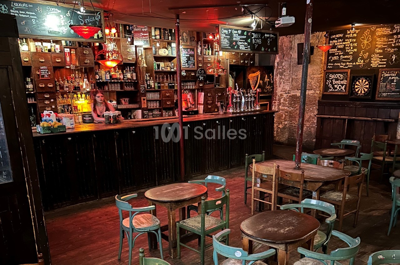 Intérieur d'un bar rustique avec tables en bois, chaises dépareillées, comptoir éclairé et ardoises affichant les menus.