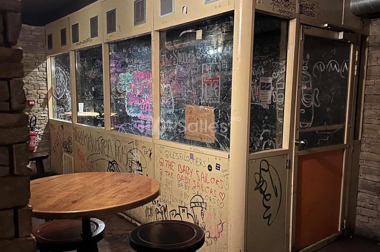 Cabine vitrée dans un bar avec des graffitis sur les murs et les vitres, entourée de tables en bois.