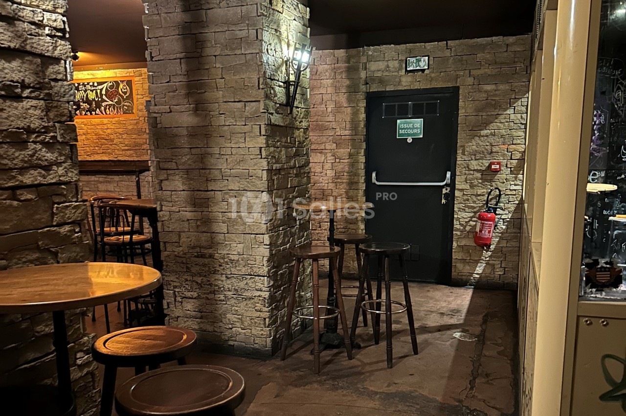 Intérieur d'un bar avec murs en pierre, tables hautes en bois, éclairage tamisé et porte de sortie de secours visible.