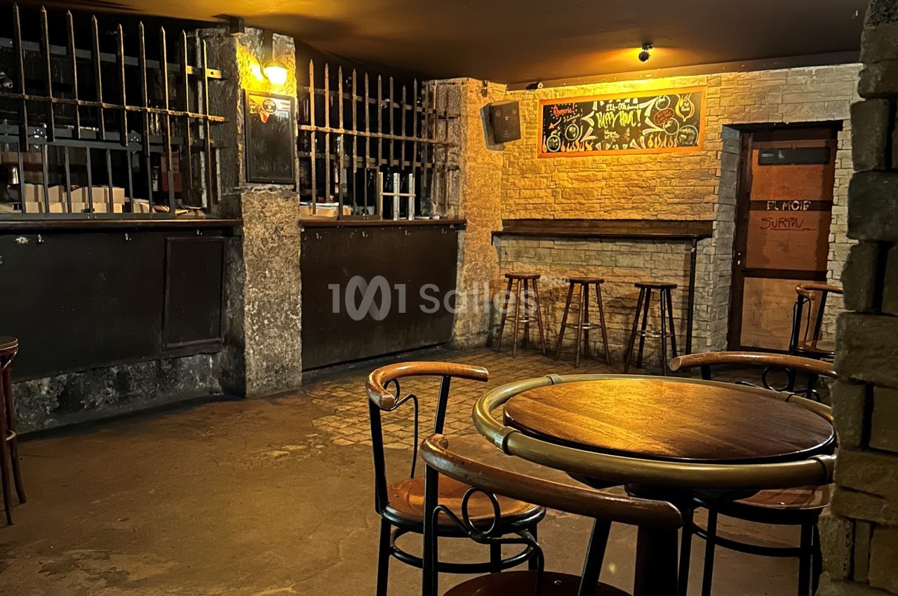 Salle de bar vide avec tables et chaises en bois, comptoir sombre, murs en briques et éclairage tamisé.