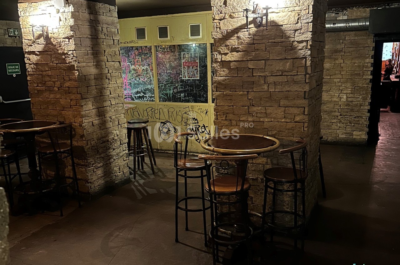 Salle avec murs en pierre, tables hautes en bois et tabourets, ambiance sombre avec graffitis sur une vitre au fond.