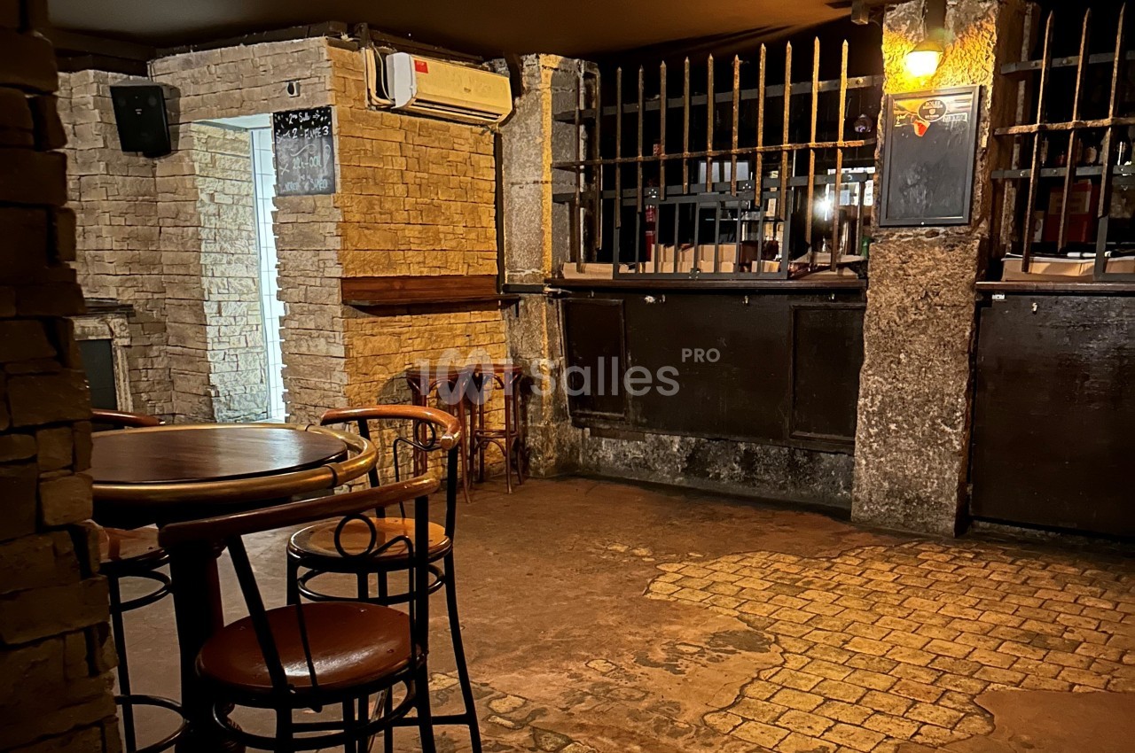 Intérieur d'un bar avec des tables en bois, des chaises hautes et un comptoir sombre derrière des barreaux métalliques.