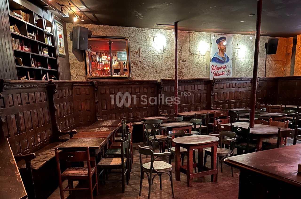 Salle de bar rustique avec tables en bois, chaises dépareillées, murs texturés et une affiche rétro accrochée.