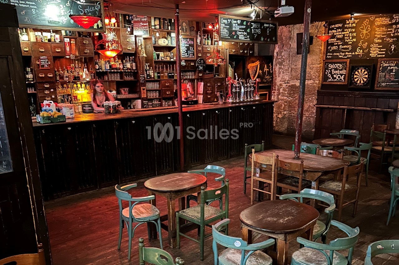 Intérieur d'un bar rustique avec tables en bois, chaises dispersées et une serveuse derrière le comptoir.