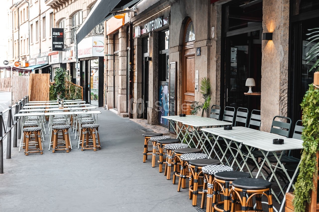 Terrasse de café avec tables et chaises alignées sur un trottoir, bordée de plantes et devantures de magasins.