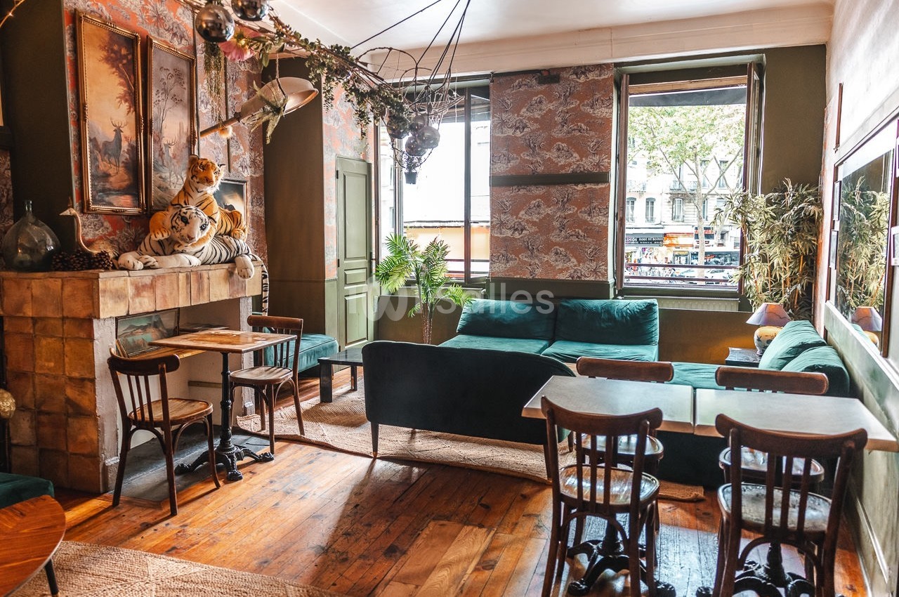 Salon lumineux avec mobilier vintage, canapé vert, tables en bois, cheminée décorative et vue sur la rue par la fenêtre.