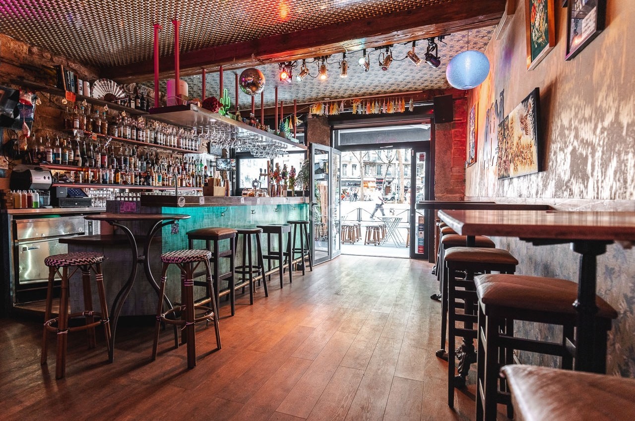 Intérieur d'un bar avec comptoir, tabourets, éclairage coloré et vue sur l'extérieur par une porte vitrée.