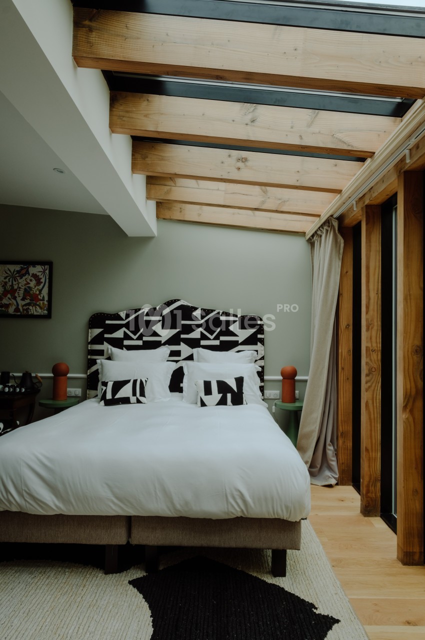 Chambre avec lit double, tête de lit à motifs géométriques, plafond en poutres apparentes et grande baie vitrée.