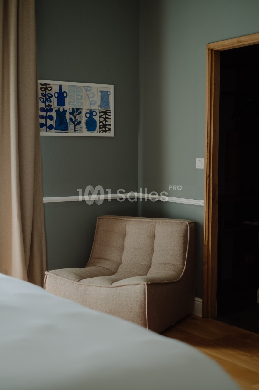 Fauteuil beige placé près d'un mur vert, avec un tableau encadré et des rideaux beiges en arrière-plan.