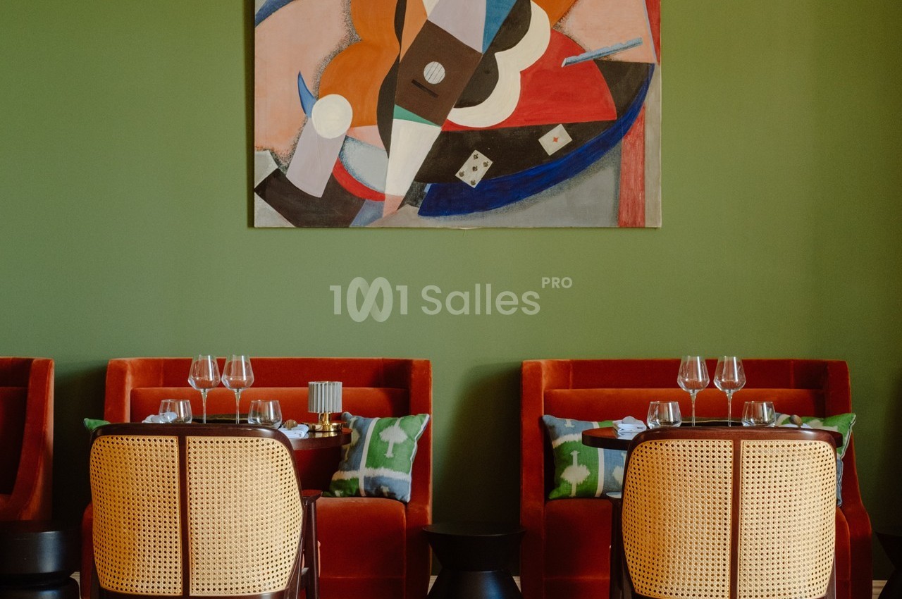 Salle de restaurant avec chaises en rotin, banquettes rouges, tables dressées et tableau abstrait sur un mur vert.