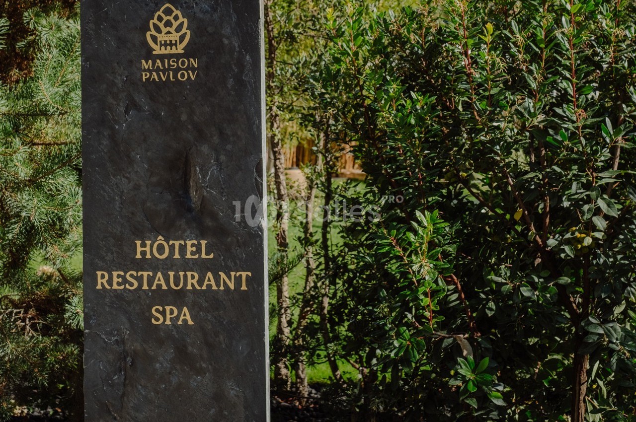 Panneau en pierre indiquant ’Hôtel Restaurant Spa Maison Pavlov’, entouré de végétation verdoyante.