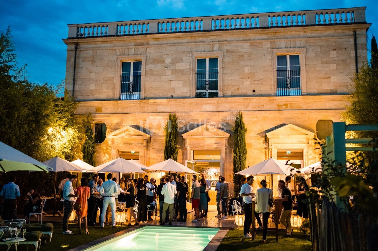 Groupe de personnes réunies en soirée dans un jardin éclairé, devant un bâtiment en pierre avec piscine au premier plan.