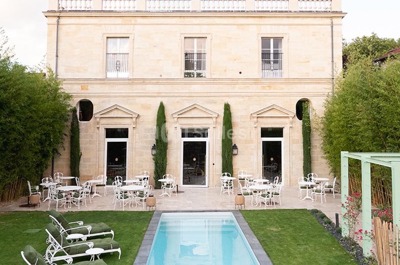 Façade d'un bâtiment en pierre avec terrasse aménagée, tables, chaises blanches et piscine au premier plan.