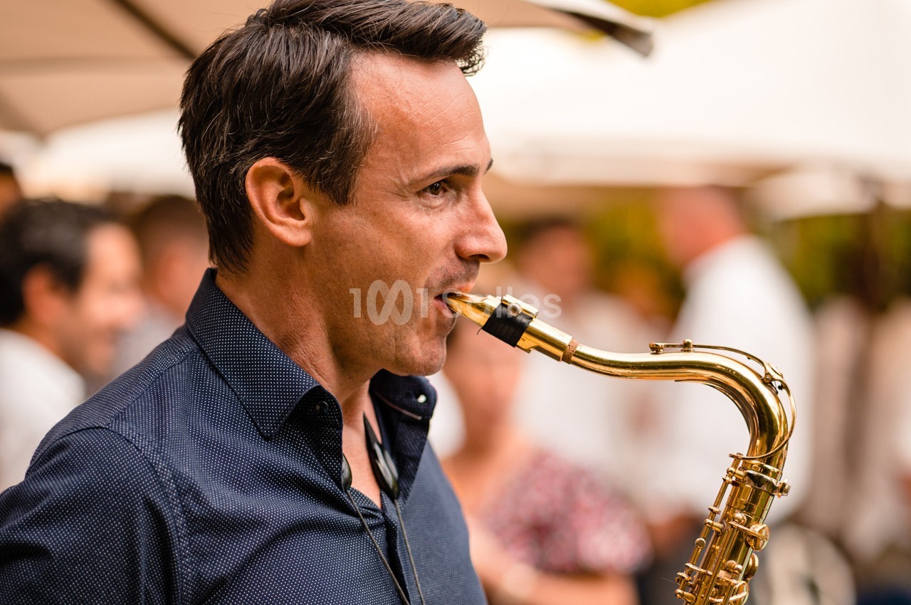 Un homme joue du saxophone en extérieur lors d'un événement, avec des parasols et des personnes floues en arrière-plan.