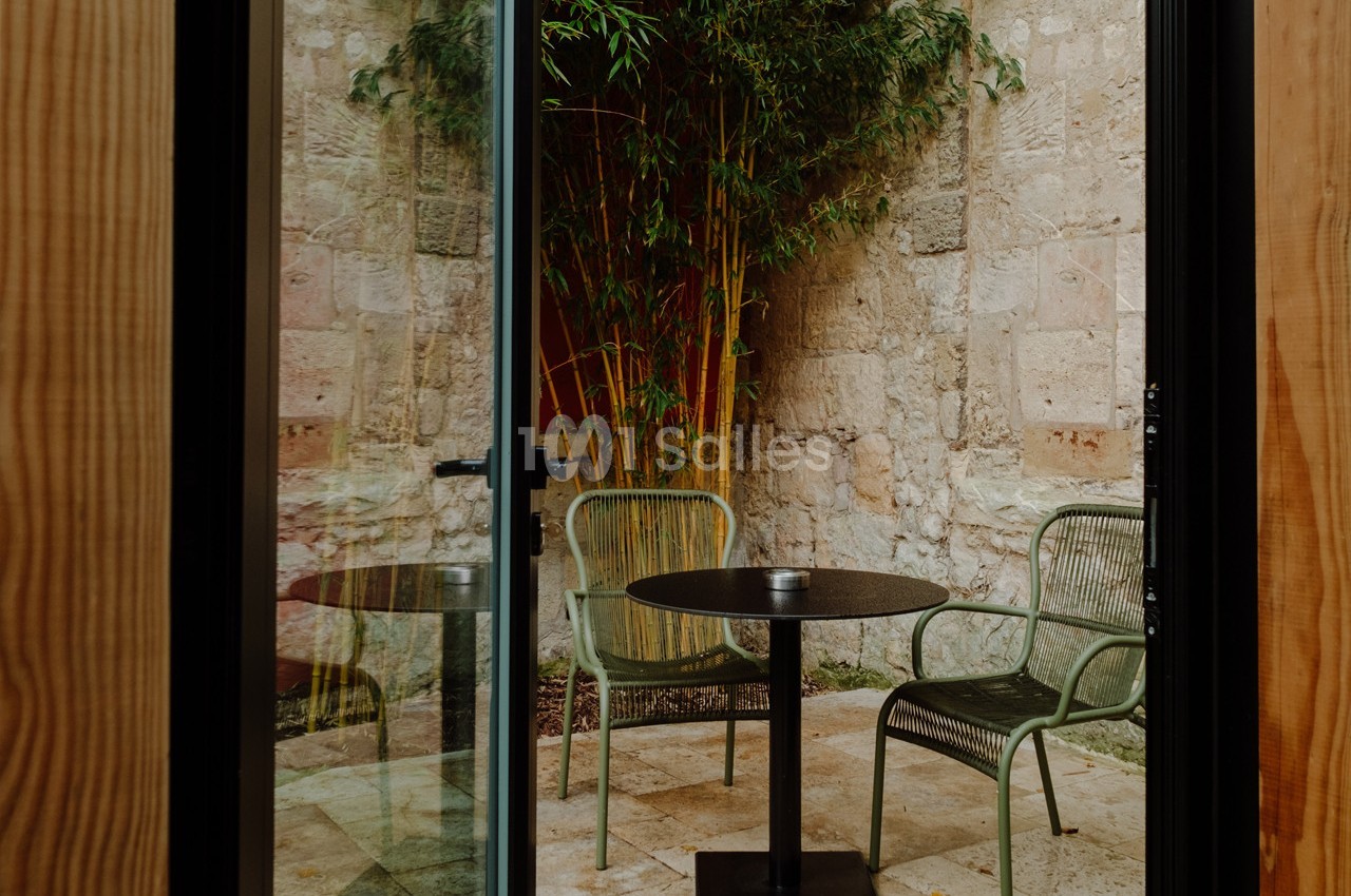 Vue sur une petite terrasse en pierre avec deux chaises vertes, une table ronde noire et des bambous en arrière-plan.