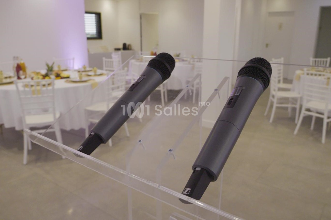 Deux microphones sans fil posés sur un pupitre transparent, avec des tables dressées en arrière-plan.