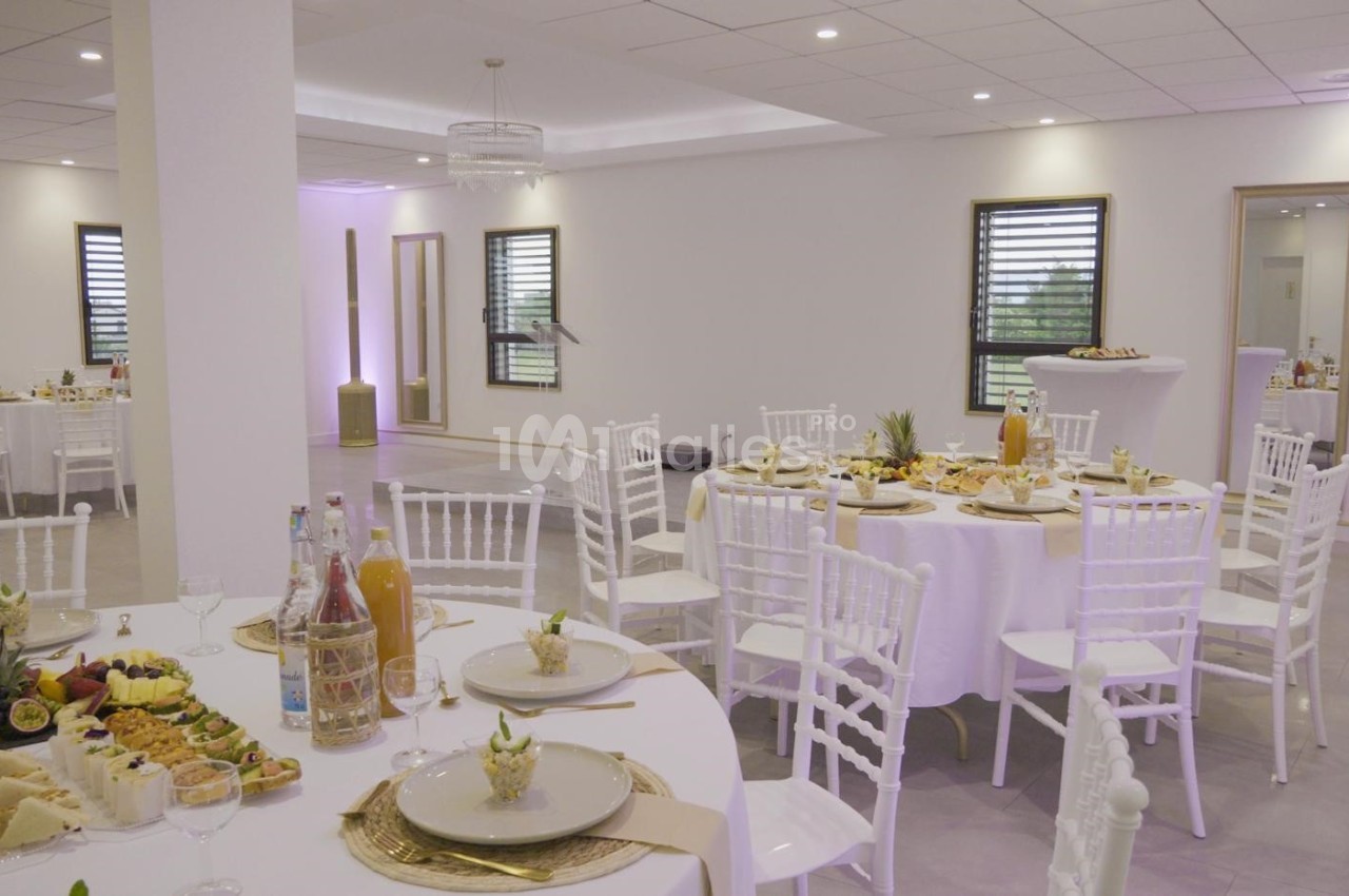 Salle de réception lumineuse avec tables rondes dressées, chaises blanches et buffet de plats variés.