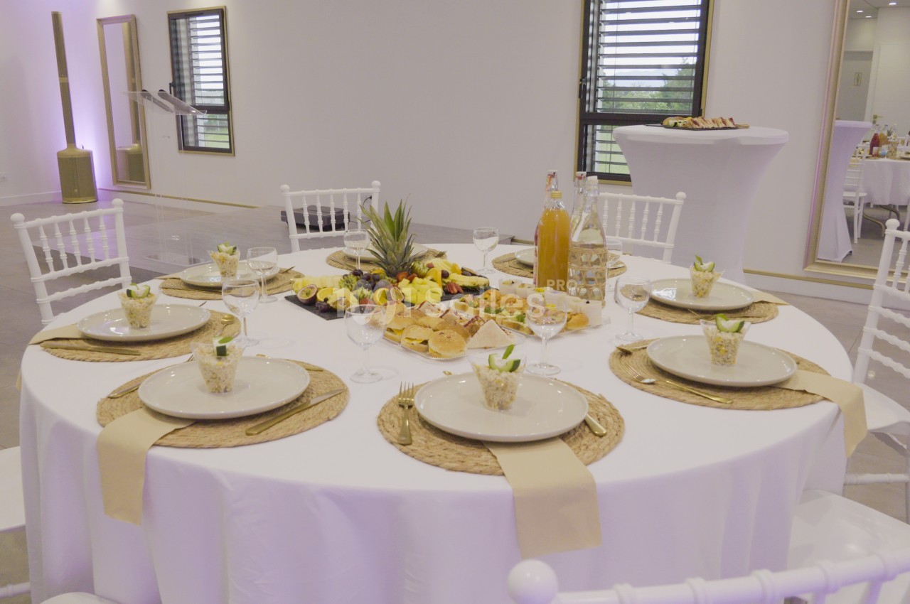 Table ronde dressée avec nappes blanches, assiettes, verres, décorations et plats variés dans une salle lumineuse.