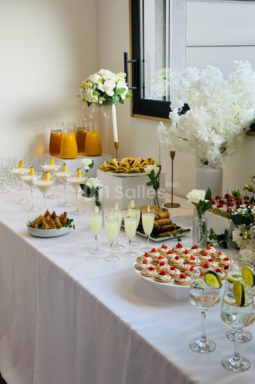 Table dressée avec des boissons, verrines, pâtisseries et décorations florales blanches dans une salle lumineuse.