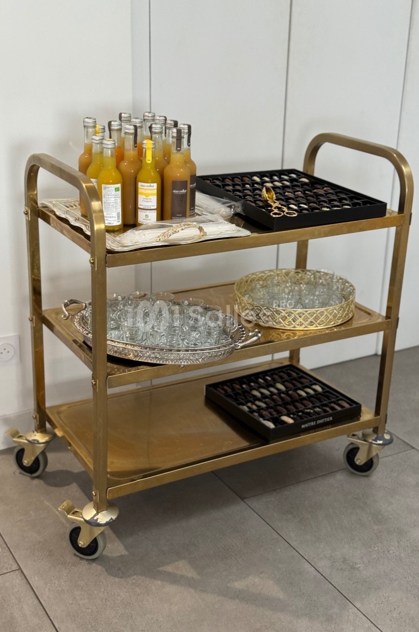 Chariot doré à trois étagères contenant des bouteilles de jus, des verres, un plateau et des chocolats.