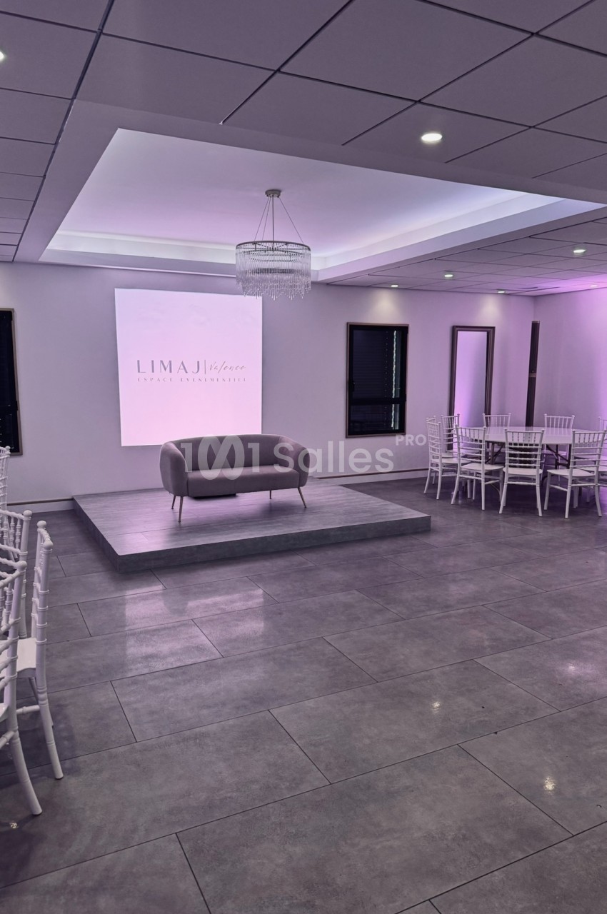 Salle de réception moderne avec éclairage violet, chaises blanches, estrade centrale et canapé gris.