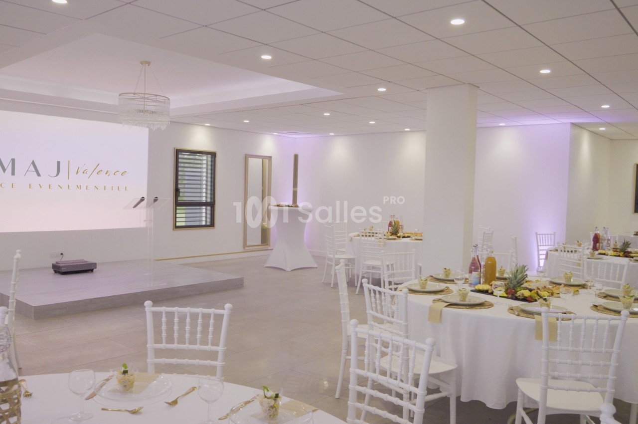 Salle de réception lumineuse avec tables rondes décorées, chaises blanches et une scène en arrière-plan.