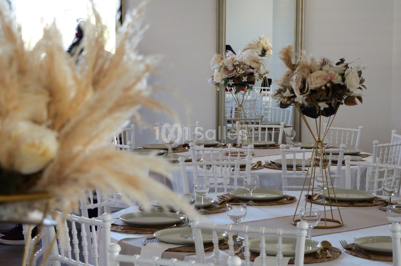 Salle de réception décorée avec des tables rondes, nappes blanches, chaises blanches et centres de table floraux.