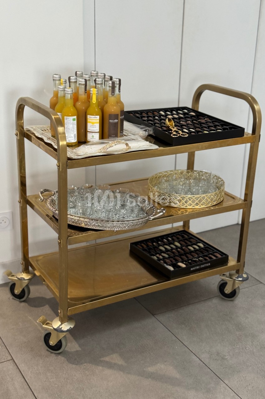 Chariot doré à trois étagères contenant des bouteilles de jus, des verres, et des plateaux de chocolats.