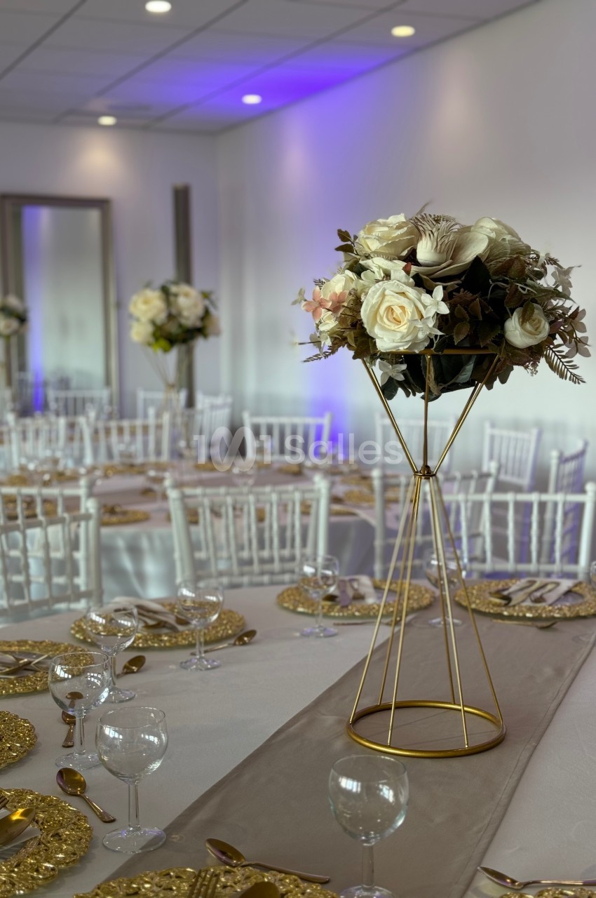 Salle de réception élégante avec tables dressées, centres de table fleuris sur supports dorés et éclairage tamisé.