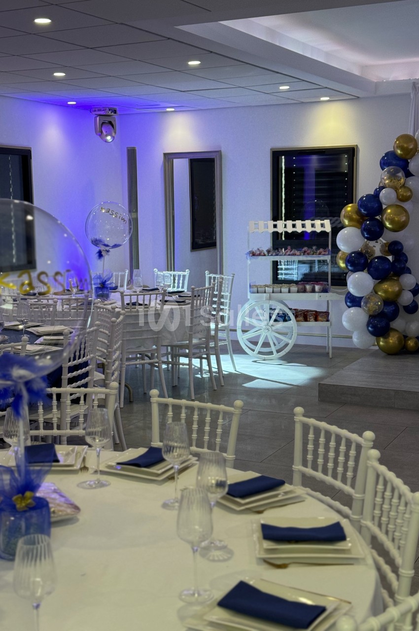 Salle de réception décorée avec des tables dressées, des ballons colorés et un chariot blanc en arrière-plan.