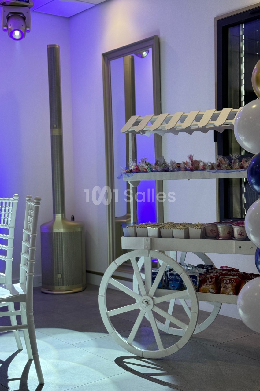 Chariot blanc décoratif avec friandises, entouré de ballons et d'une chaise blanche dans une salle éclairée en bleu.