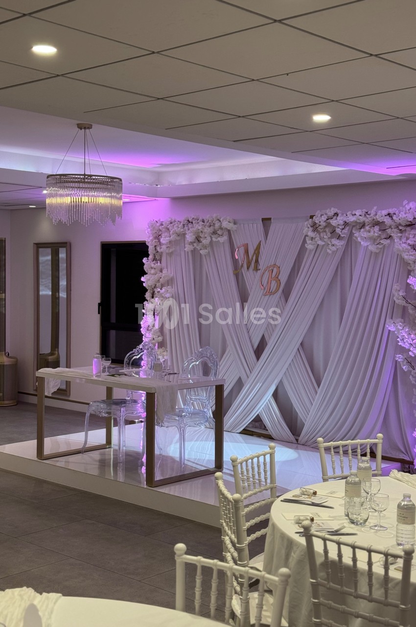 Salle de réception décorée avec des rideaux blancs, des fleurs, des chaises blanches et un éclairage rose.