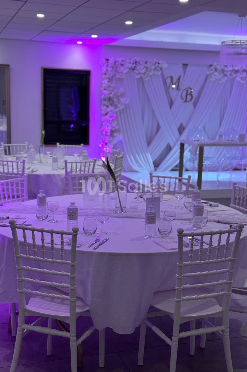 Salle de réception décorée avec des tables rondes blanches, des chaises élégantes et un fond lumineux violet.
