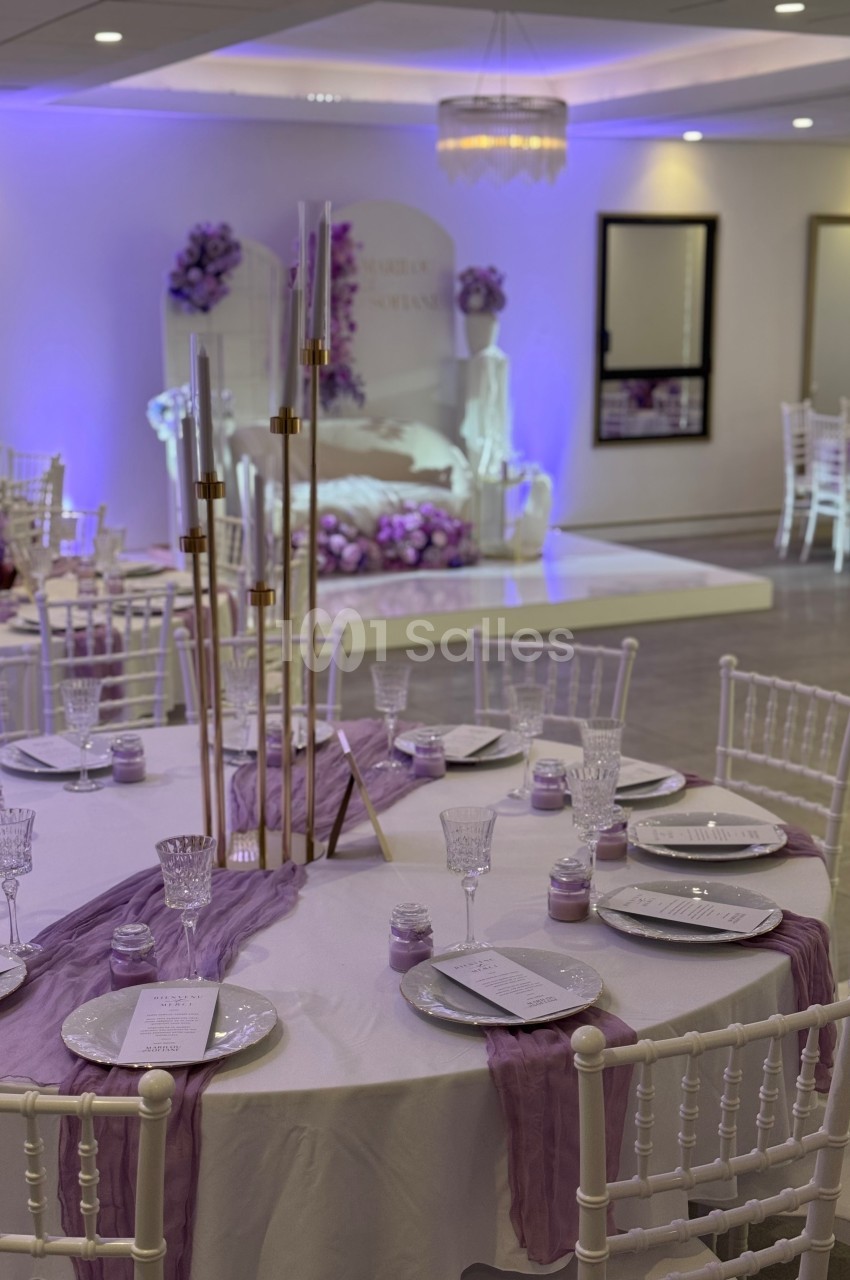 Salle de réception décorée en violet avec tables dressées, chaises blanches et éclairage tamisé.