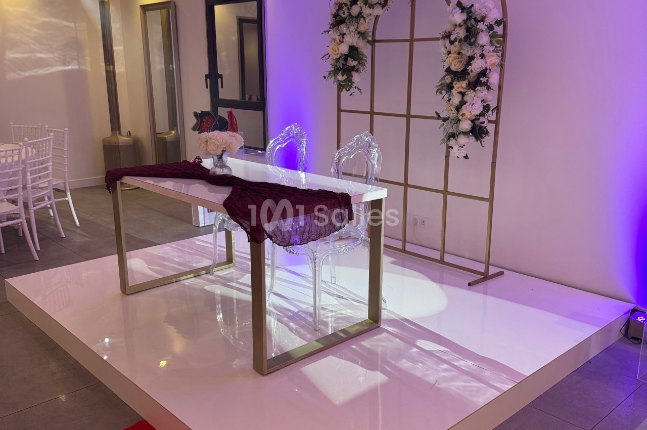 Table blanche décorée d'un tissu bordeaux, deux chaises transparentes et une arche florale dans une salle éclairée.