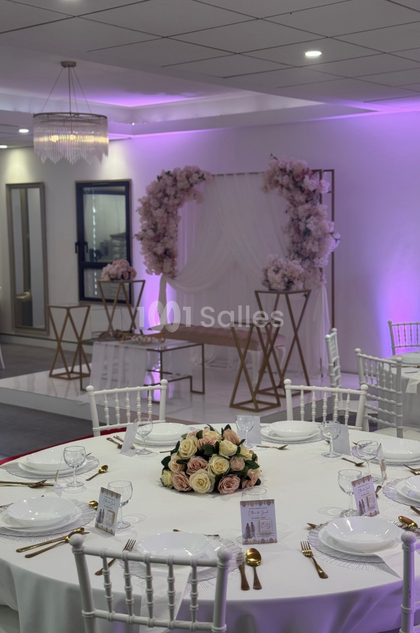 Salle de réception décorée avec des tables dressées, un fond floral rose et blanc, et un éclairage violet.