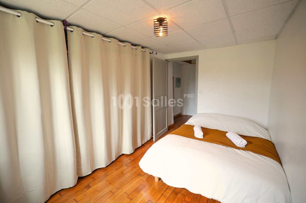 Chambre lumineuse avec un lit double, draps blancs, plaid ocre, rideaux beiges et parquet en bois.