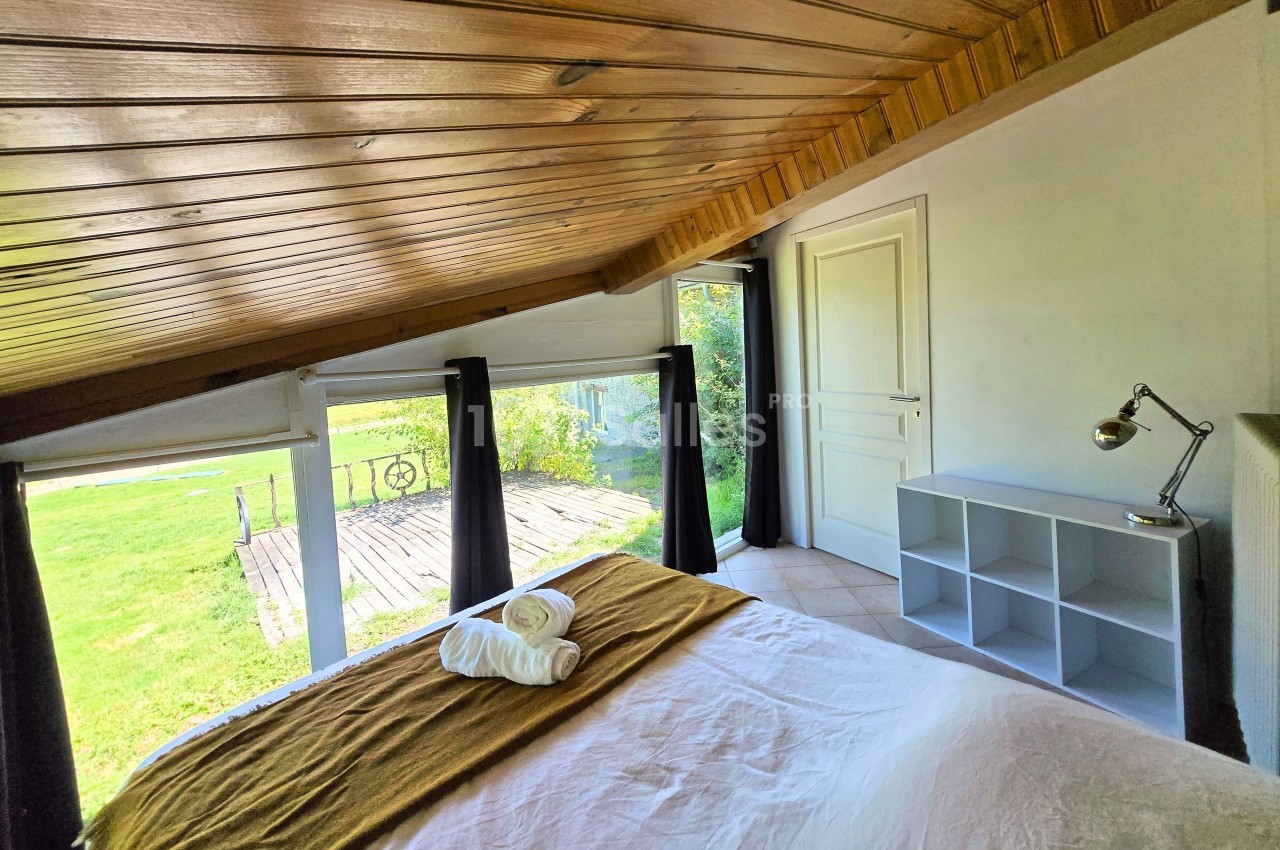 Chambre lumineuse sous combles avec lit double, étagère, lampe de bureau et vue sur un jardin par de grandes fenêtres.