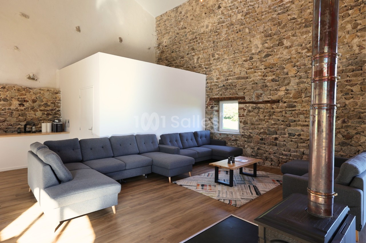 Salon spacieux avec murs en pierre, canapé d'angle gris, poêle à bois et lumière naturelle provenant d'une fenêtre.