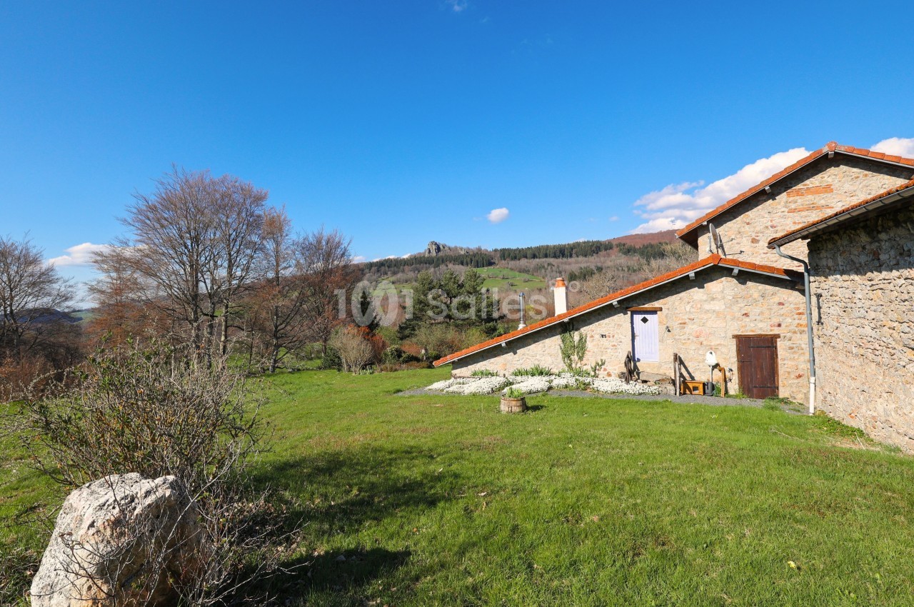 Maison en pierre avec toit en tuiles, entourée de verdure et de collines sous un ciel bleu dégagé.