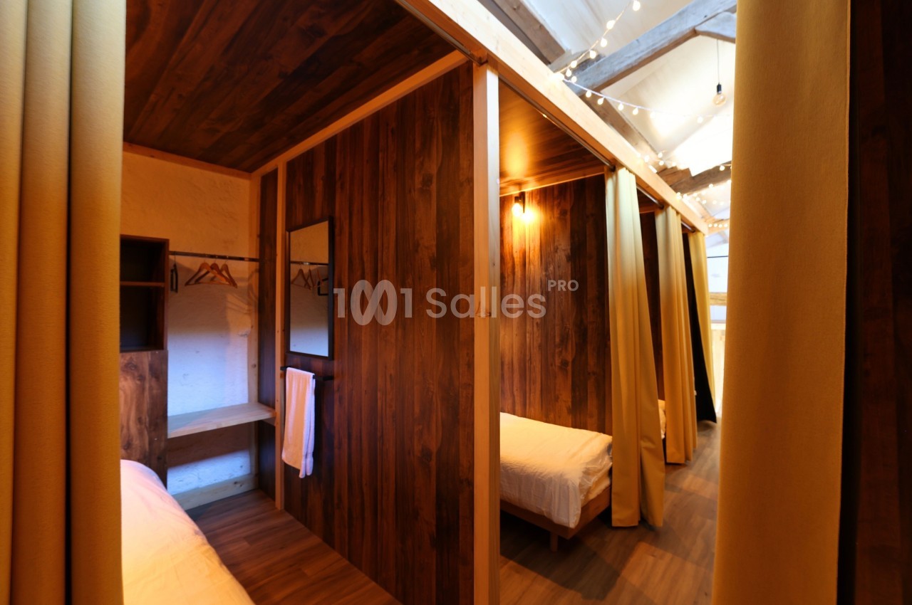 Cloisons en bois séparant des lits simples dans un dortoir avec rideaux, éclairage chaleureux et rangements intégrés.
