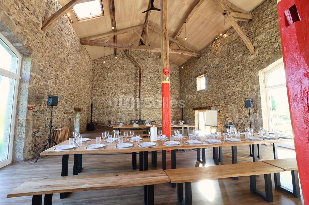 Salle en pierre avec poutres apparentes, tables en bois dressées avec vaisselle, éclairée par des fenêtres et un puits de…