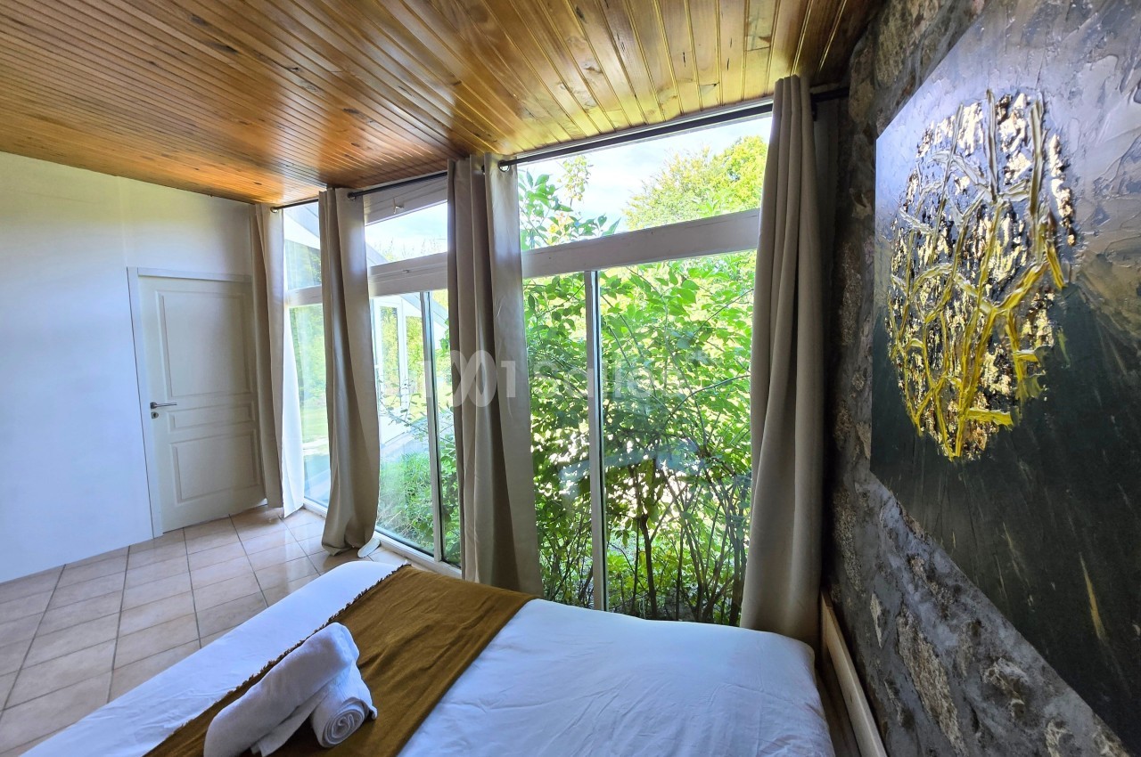 Chambre lumineuse avec grande baie vitrée donnant sur un jardin verdoyant, plafond en bois et lit avec linge plié.