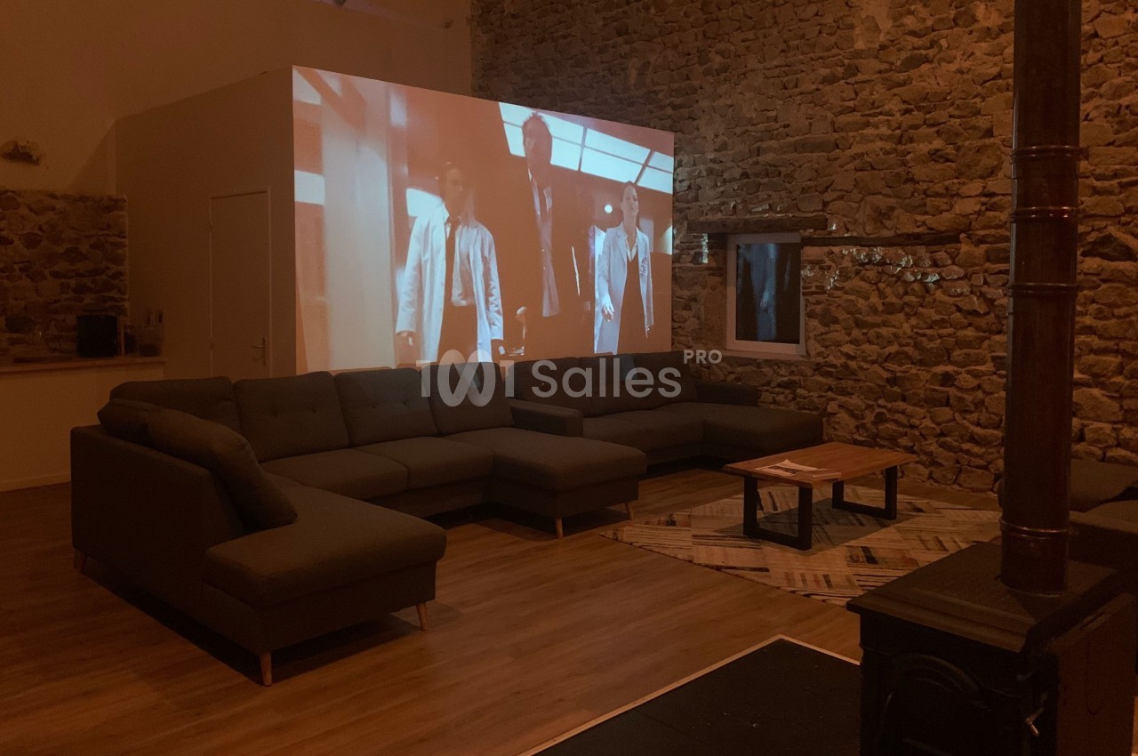 Projection d'un film sur un mur en pierre dans un salon avec canapé d'angle, table basse et poêle à bois.