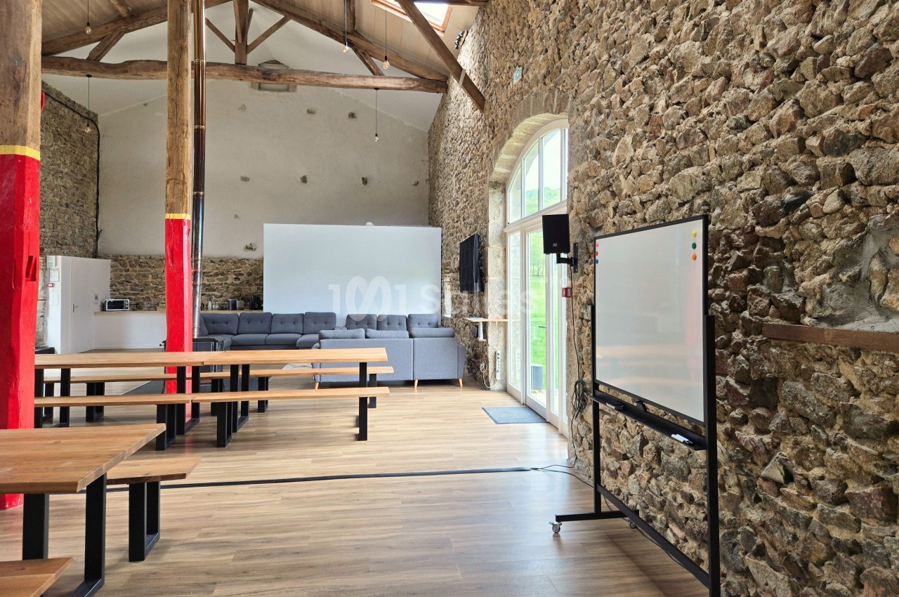 Salle lumineuse avec murs en pierre, grandes fenêtres, tables en bois, tableau blanc et canapés gris au fond.