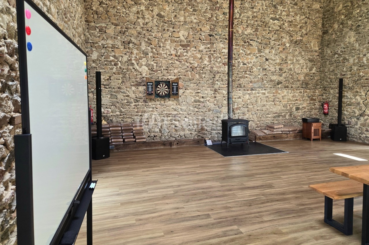 Salle avec murs en pierre, tableau blanc, poêle à bois, cible de fléchettes et bancs en bois sur un sol en parquet.