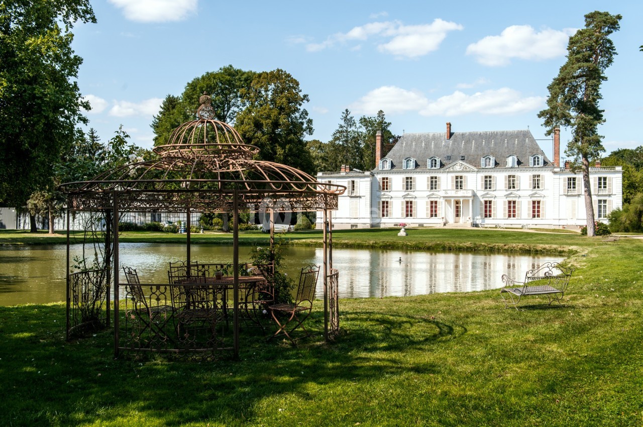 Pavillon en fer forgé près d'un étang, avec un manoir blanc en arrière-plan entouré de verdure.