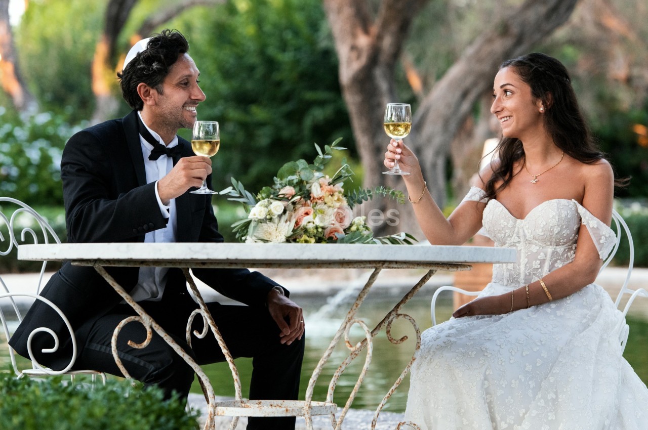 Un couple en tenue de mariage trinque avec des verres de vin blanc, assis à une table en extérieur.