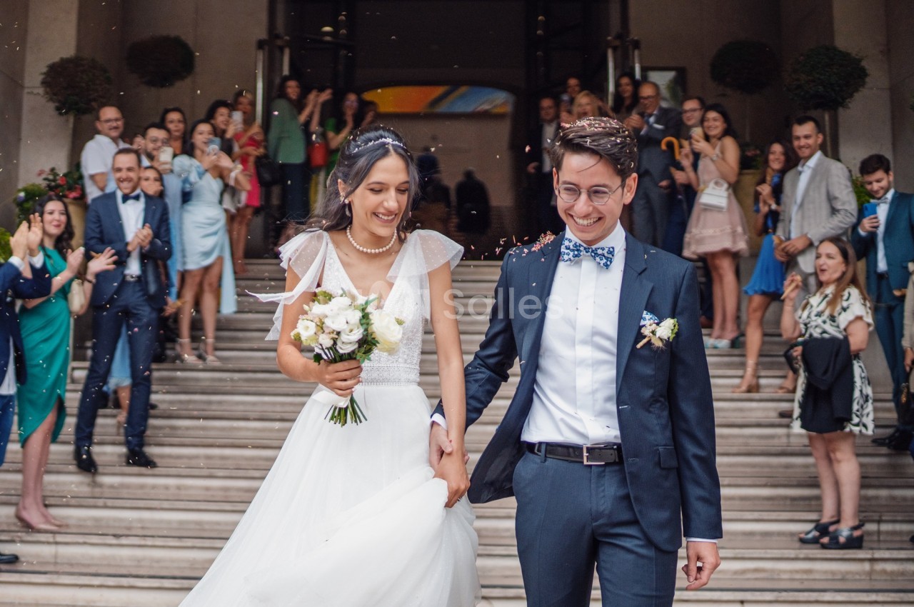 Un couple souriant sort d'un bâtiment après leur mariage, entouré d'invités applaudissant sur les marches.