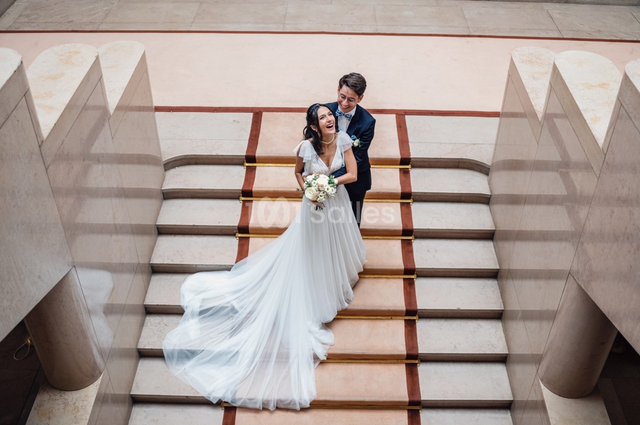 Un couple de mariés souriant pose sur un escalier en pierre, la mariée tenant un bouquet de fleurs.
